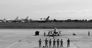 Black and white image of military personnel and jet at Istanbul airfield with parked aircraft in the background.